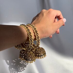 May include: A pair of gold bangles with pearl accents and decorative jingles. The bangles are worn on a person's wrist.