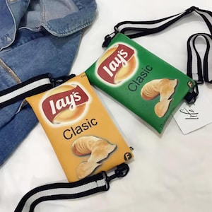 May include: Two novelty shoulder bags designed to resemble Lay's Classic potato chip bags. One bag is yellow, the other green, both with the Lay's logo and chip graphics. Each bag has a black and white striped strap.