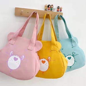 May include: Three canvas tote bags in pink, yellow, and light blue, each featuring a bear-shaped silhouette with rounded ears and a printed character face on the front. The bags have matching fabric handles and are hanging from a wooden wall rack.