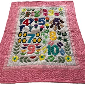 May include: A pink quilted baby blanket with a white center featuring embroidered numbers 1 through 10, various animals, and flowers.