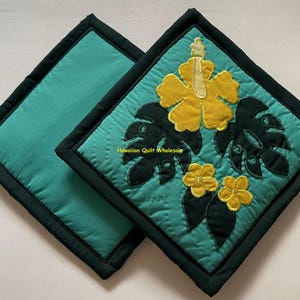 May include: Two square teal pot holders with dark green trim. One pot holder features a yellow hibiscus flower and dark green leaves. The other pot holder is solid teal. The text "Hawaiian Quilt Wholesale" is visible.
