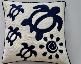 May include: Square decorative pillow with a quilted off-white background and navy blue sea turtle and sun designs. The pillow has a navy blue border and a zipper closure.