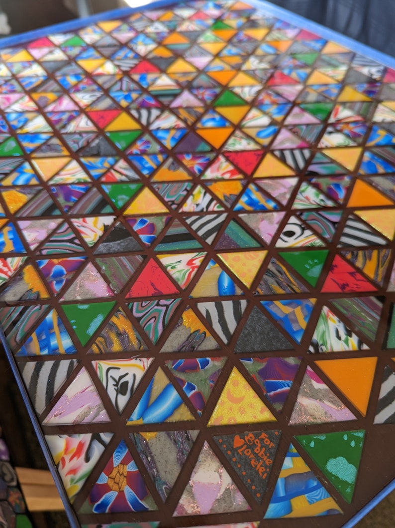 May include: A colorful mosaic table top with a geometric pattern of triangles. The table top is made of various materials, including wood, glass, and resin. The table top is decorated with a heart and the words "For Bobbie Love!" in orange.