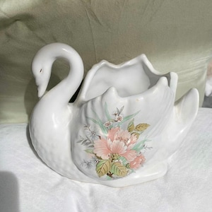 Puede incluir: Macetero de cerámica blanca en forma de cisne con un diseño floral con flores rosas y verdes. El cuello del cisne está curvado y el macetero tiene una abertura ancha.
