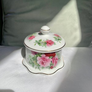 May include: A white ceramic trinket box with a lid featuring a floral design of pink and red roses with green leaves. The box has a gold trim around the top and bottom.