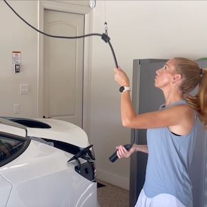 May include: A woman in a light blue tank top and white shorts is holding a black electric car charging cable. The cable is attached to a white electric car. The car is parked in a garage.