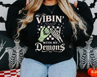 May include: Black sweatshirt with the text "VIBIN' WITH MY DEMONS" and a graphic of a green hand and a skeleton hand pinky-promising. The design includes bats and stars. The model is wearing blue jeans.