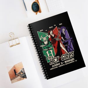May include: A black spiral notebook with a colorful illustration of a band called "The Hex Girls". The illustration features three women in gothic attire playing musical instruments. The text "October 31 * midnight" and "salem arena * oakhaven, massachusetts * 18+" is printed below the illustration.
