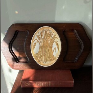 Op de afbeelding: Een vintage houten serveerblad met een tarwearen-ontwerp. De lade heeft een donkerbruine afwerking met uitgesneden handgrepen. Het midden heeft een ovale keramische tegel met een gedetailleerde illustratie van tarwearen.