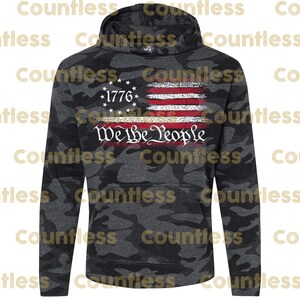 May include: A black and grey camouflage hoodie with a distressed American flag design and the text "We The People" printed on the front. The flag is red, white and blue with a gold star in the top left corner. The text "1776" is printed above the flag.