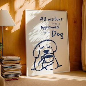 Puede incluir: Un cuadro enmarcado con el texto "All visitors must be approved by the Dog". La obra presenta un dibujo lineal azul de un perro sosteniendo una taza. El cuadro está en un marco blanco y se apoya en una pared de madera.