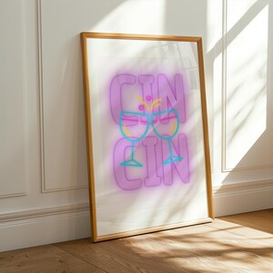 May include: A framed print featuring the word "GIN" in large, purple, neon-style letters. Two cocktail glasses are depicted in neon yellow, pink, and blue. The artwork is leaning against a white wall and a wooden floor.