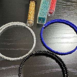May include: An overhead shot features jewelry-making supplies. There are four beaded necklaces in clear, black, and blue. Three tubes contain gold, teal, and red beads. The beads are small and round, perfect for crafting.