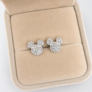 May include: A pair of silver-colored stud earrings in the shape of a mouse head, encrusted with small, sparkling crystals. The earrings are displayed inside a beige jewelry box with a soft, velvety interior. The earrings are a classic and playful accessory.