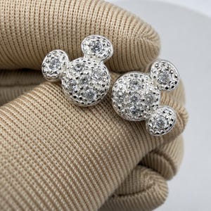 May include: A pair of silver-colored stud earrings in the shape of a mouse head. The earrings are covered in small, sparkling clear stones. The earrings are displayed on a tan fabric surface.