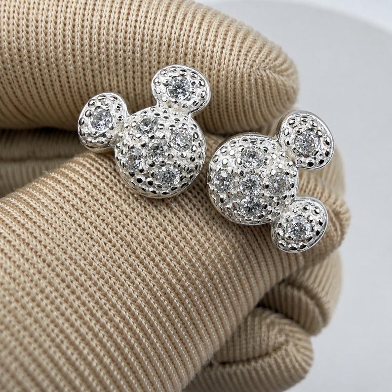 May include: A pair of silver-colored stud earrings in the shape of a mouse head. The earrings are covered in small, sparkling clear stones. The earrings are displayed on a tan fabric surface.