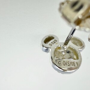 May include: Close-up of a silver-colored Mickey Mouse-shaped earring. The earring has three circular elements, with the center one bearing the "&copy;DISNEY" mark. The earring is marked "S925".