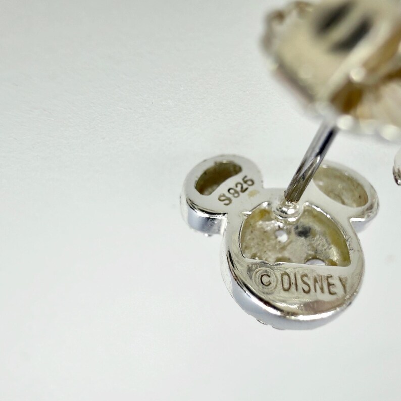 May include: Close-up of a silver-colored Mickey Mouse-shaped earring. The earring has three circular elements, with the center one bearing the "&copy;DISNEY" mark. The earring is marked "S925".