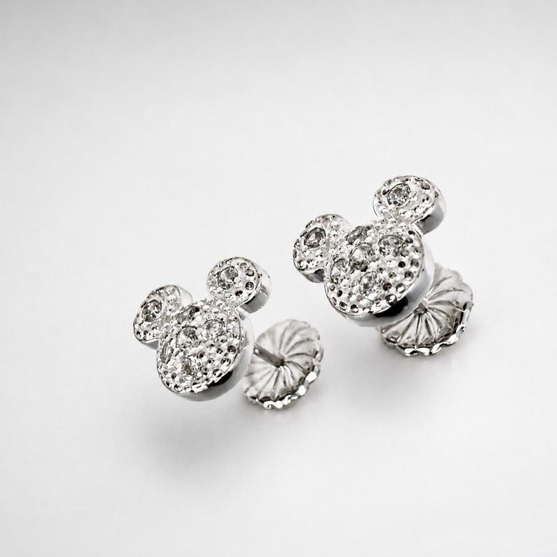May include: A pair of silver-toned stud earrings in the shape of Mickey Mouse's head. Each earring is encrusted with small, sparkling clear stones. The earrings are displayed on a white background.