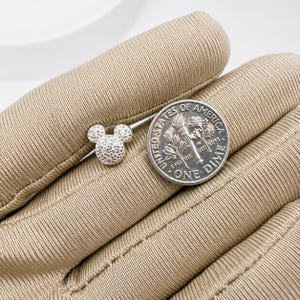 May include: A small, silver-colored stud earring in the shape of Mickey Mouse's head, encrusted with small, sparkling stones. A U.S. dime is next to the earring for size comparison. The earring is on a tan surface.