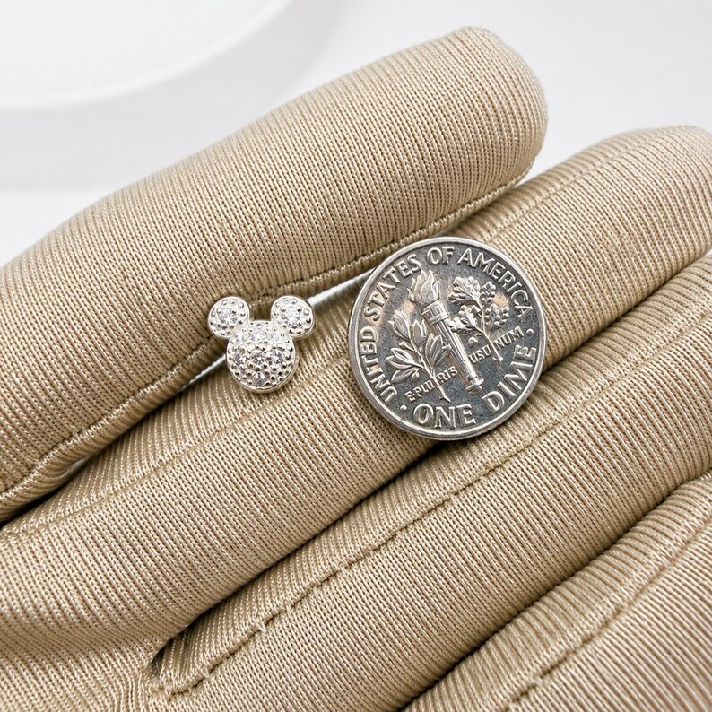 May include: A small, silver-colored stud earring in the shape of Mickey Mouse's head, encrusted with small, sparkling stones. A U.S. dime is next to the earring for size comparison. The earring is on a tan surface.