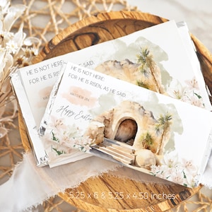 Könnte beinhalten: Zwei weiße Schokoladenriegel-Verpackungen mit einer Aquarell-Illustration eines Steinbogens, Palmen und weißen Blumen. Der Text auf den Verpackungen lautet "Er ist nicht hier: Denn er ist auferstanden, wie er gesagt hat. Matthäus 28,6" und "Frohe Ostern".