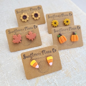 May include: A collection of handmade earrings displayed on brown cards. The earrings feature fall-themed designs, including sunflowers, maple leaves, pumpkins, and candy corn. The cards are labeled "Sunflower Place Co."