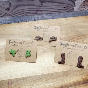 May include: Three pairs of handmade earrings on display. One pair features green cactus shapes, another has brown cowboy hat designs, and the last showcases brown cowboy boots. Each pair is mounted on a card with the text "Sunflower Place Co."