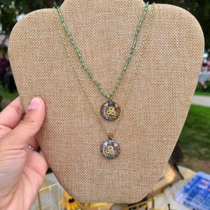May include: A gold chain necklace with two pendants. The pendants are round and feature a floral design with blue, white, and orange colors. The necklace is displayed on a brown fabric mannequin.