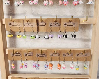 May include: A wooden display rack showcasing various pairs of dangle earrings. The earrings feature cartoon character designs in a variety of colors, including pink, white, black, and yellow. The earrings are displayed on small brown cards.