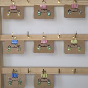 Könnte beinhalten: Ein Holzregal mit mehreren Paaren von ausgefallenen Ohrringen. Jedes Paar ist mit einer Büroklammer an einer kleinen braunen Karte befestigt. Die Ohrringe zeigen verschiedene Cartoon-Frosch-Designs in Grün, Weiß und Schwarz.