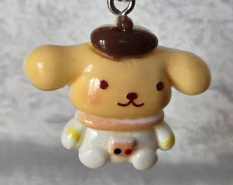 May include: A yellow cartoon dog charm with a brown beret. The charm has a white outfit with a pink collar and red and black accents. The dog has red cheeks and a black nose and mouth. The charm is suspended from a silver-colored metal loop.