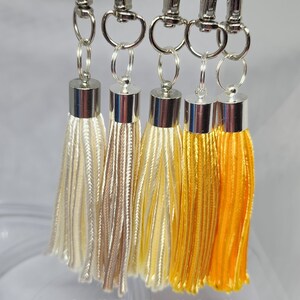 May include: Five tassel keychains with silver clasps. The tassels are in shades of white, beige, and yellow. The numbers 4, 25, 3, 26, and 2 are printed below the tassels.