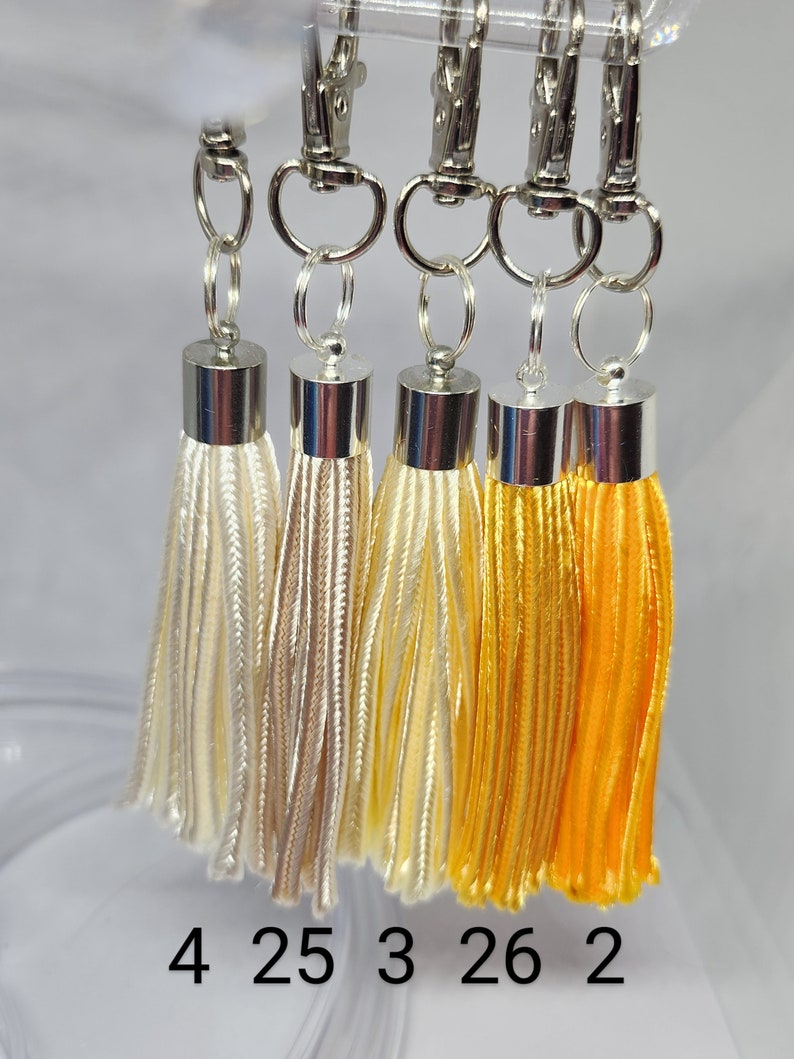 May include: Five tassel keychains with silver clasps. The tassels are in shades of white, beige, and yellow. The numbers 4, 25, 3, 26, and 2 are printed below the tassels.
