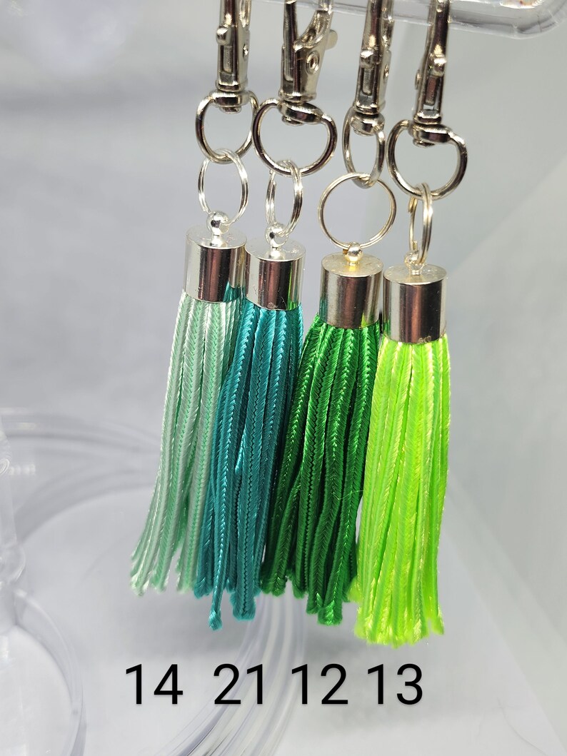 May include: Four green tassel keychains with silver clasps and rings. The tassels are different shades of green, from light green to dark green. The numbers 14, 21, 12, and 13 are printed below the tassels.