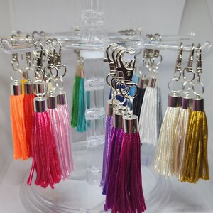 May include: A display of colorful tassel keychains with silver key rings. The tassels are made of various colors, including orange, pink, white, purple, and gold.
