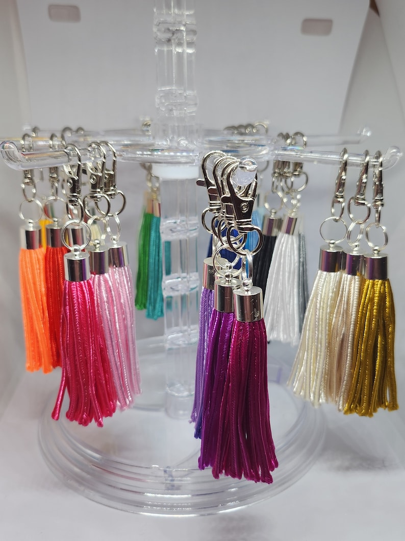 May include: A display of colorful tassel keychains with silver key rings. The tassels are made of various colors, including orange, pink, white, purple, and gold.