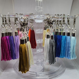 May include: A display of colorful tassel keychains with silver key rings. The tassels are in various colors, including purple, lavender, gold, white, gray, blue, and light blue.