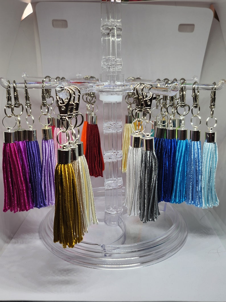 May include: A display of colorful tassel keychains with silver key rings. The tassels are in various colors, including purple, lavender, gold, white, gray, blue, and light blue.