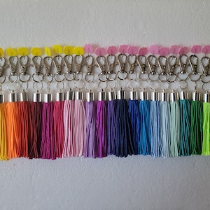 Könnte beinhalten: Ein Set mit 24 bunten Quasten-Schlüsselanhängern mit silbernen Metallverschlüssen. Die Quasten sind aus weichem Garn gefertigt und in verschiedenen Farben erhältlich, darunter Weiß, Gelb, Orange, Rot, Rosa, Lila, Blau, Grün und Schwarz.
