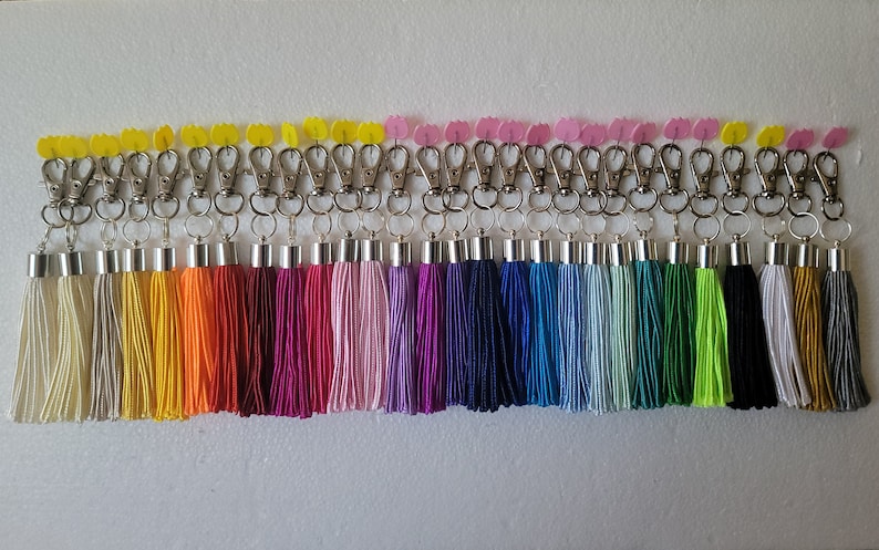 May include: A set of 24 colorful tassel keychains with silver metal clasps. The tassels are made of soft yarn and come in a variety of colors, including white, yellow, orange, red, pink, purple, blue, green, and black.