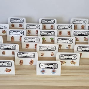 Pode incluir: Exibição de vários brincos de pressão em cartões brancos, dispostos em um expositor de madeira em camadas. Os brincos apresentam diferentes designs, incluindo ursinhos de pelúcia, animais e outras pequenas figuras. Os cartões têm um logotipo e a frase "HotHandbags & Accessories".
