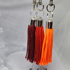 May include: Three tassel keychains with silver clasps. The tassels are red, orange, and a darker shade of red. The keychains are hanging from a clear plastic display stand. The numbers 5, 6, and 7 are printed on the stand.