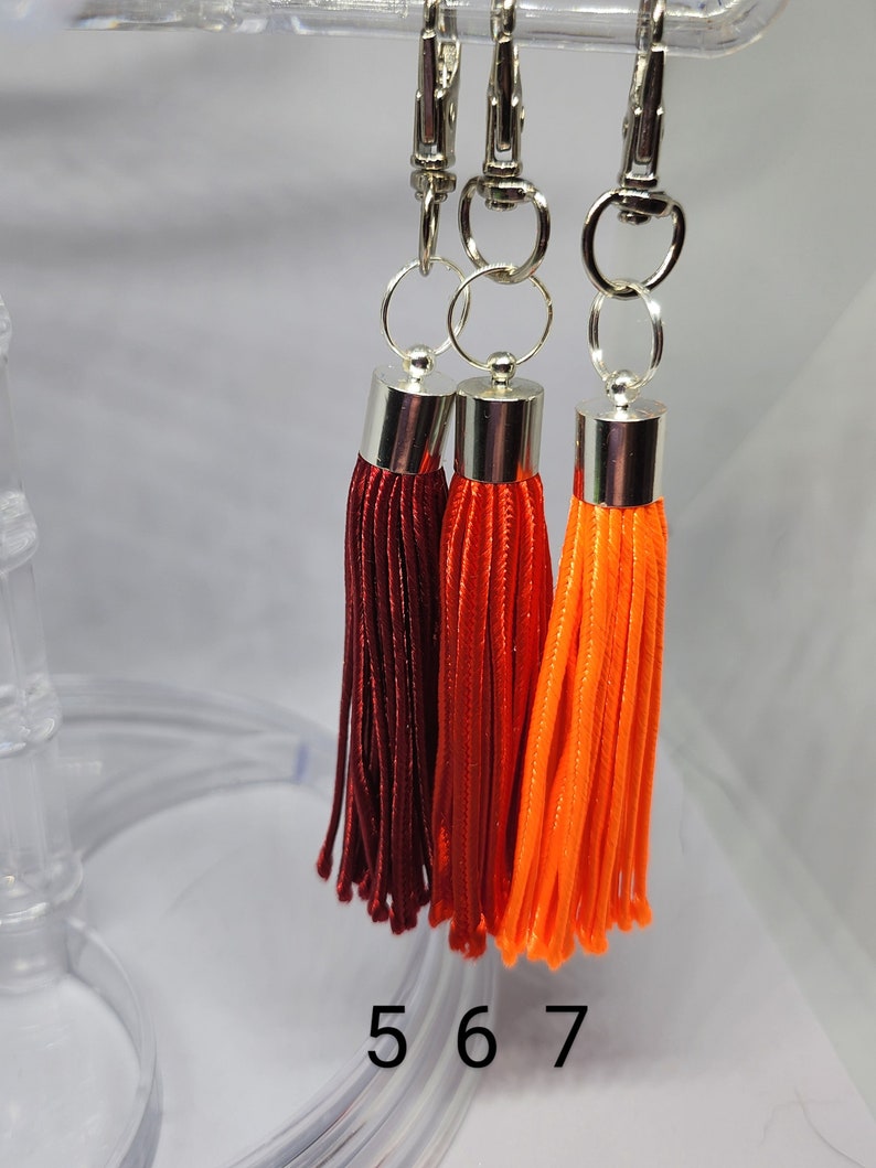 May include: Three tassel keychains with silver clasps. The tassels are red, orange, and a darker shade of red. The keychains are hanging from a clear plastic display stand. The numbers 5, 6, and 7 are printed on the stand.