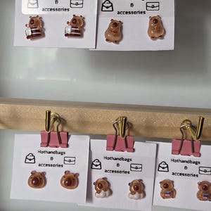 May include: Three cards with pink clips hanging from a wooden bar. Each card features a different design of brown bear earrings with the text "Hothandbags 8 accessories" on each card.