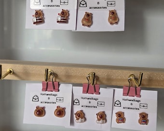 May include: Three cards with pink clips hanging from a wooden bar. Each card features a different design of brown bear earrings with the text "Hothandbags 8 accessories" on each card.
