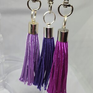 May include: Three purple and lavender tassel keychains with silver clasps and rings. The tassels are different shades of purple, with the numbers 8, 22, and 9 printed below them.