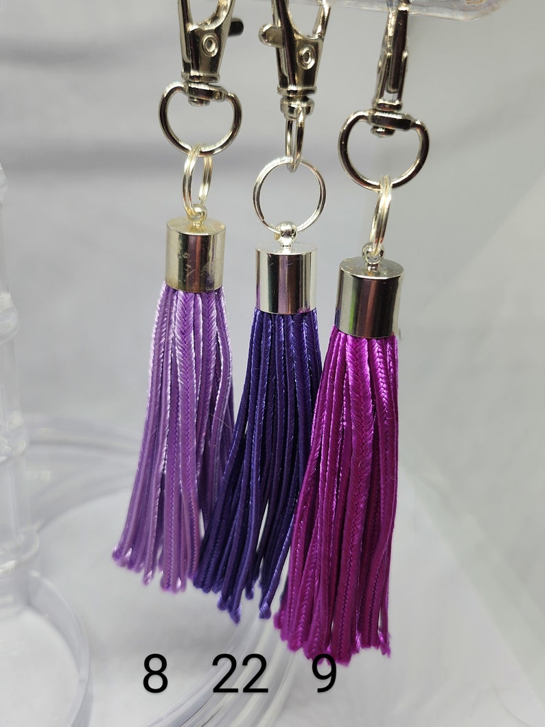 May include: Three purple and lavender tassel keychains with silver clasps and rings. The tassels are different shades of purple, with the numbers 8, 22, and 9 printed below them.