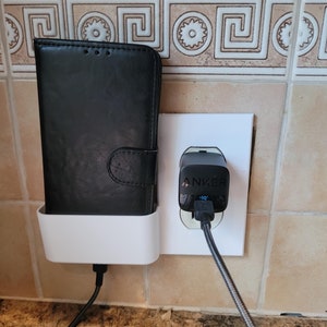 May include: A black phone case is plugged into a white wall socket with a black Anker charger. The phone case is in a white phone holder.