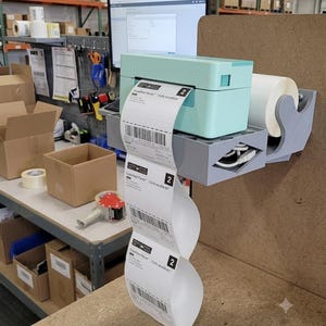 Puede incluir: Una impresora térmica de etiquetas en color verde menta claro sobre un soporte gris impreso en 3D fijado a un tablero de madera. La impresora sostiene un rollo de etiquetas blancas, con una tira de etiquetas de envío con códigos de barras.